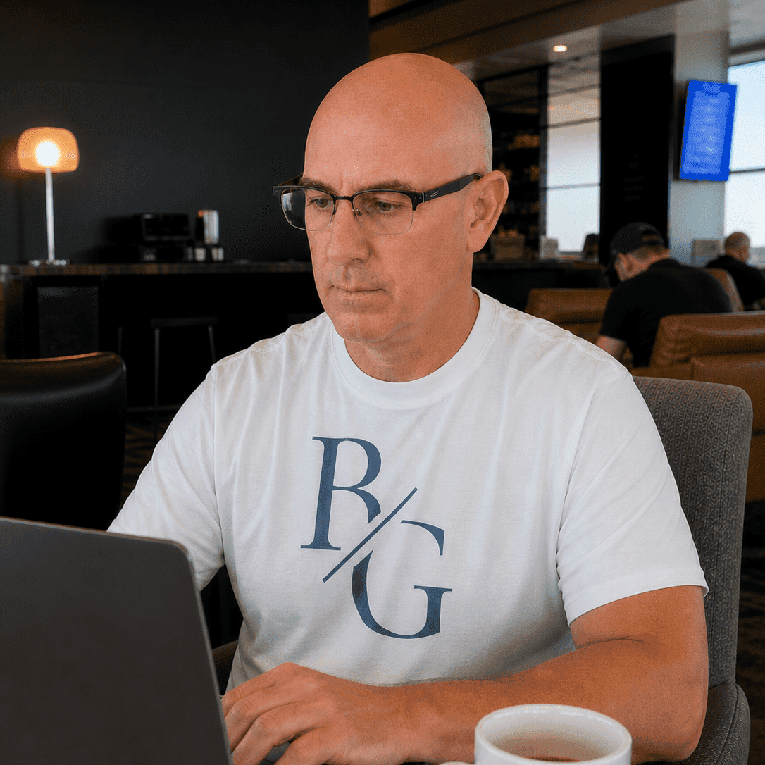 Brian Gibbs working on a laptop in an airport lounge, wearing a white BG monogram t-shirt with a coffee cup on the table